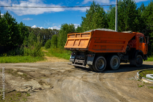Bild auf Leinwand Orange dumper truck loaded with gravel standing on dirt road side in sunny weather