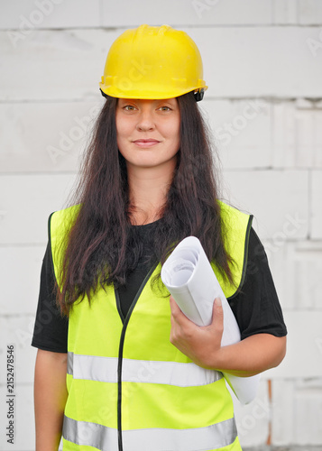 Young female construction worker in hi vis vest, helmet and gloves, carrying cable roll on her shoulder, bare new house wall near