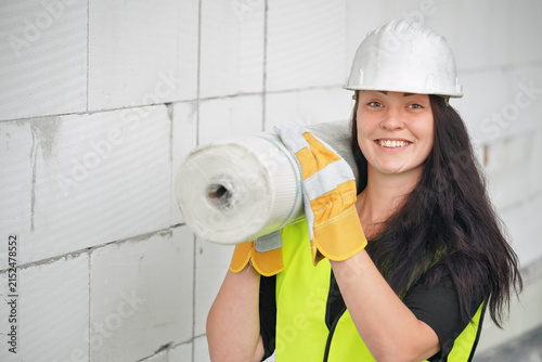 Young woman in green high visibility vest and helmet carrying roll with construction material on her shoulder near build site wall