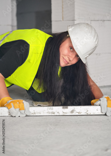 Construction worker at new house building site, kneeling, spirit level on ground in front of her, looking into camera, bare wall behind