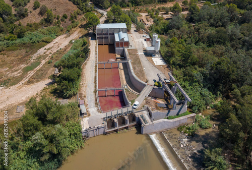 Top-down view of a gated river intake facility with potassium permanganate-dosed red water channels, concrete sedimentation basins, storage silos and processing buildings surrounded by trees