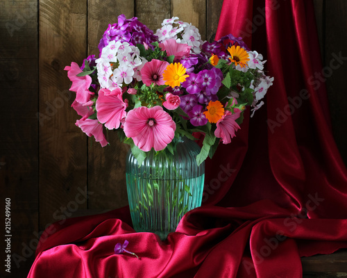 Colorful lavatera and phlox bouquet in glass vase with red satin fabric and rustic wooden background