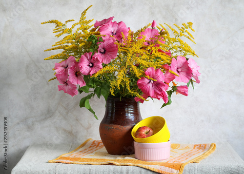 Bouquet of pink lavatera and goldenrod flowers in ceramic vase with apple and colorful bowls on table
