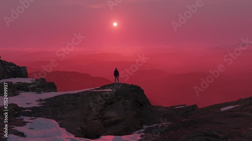 Lone Figure on Snowy Mountain Peak Gazing at a Surreal Red Sky Sunset Landscape