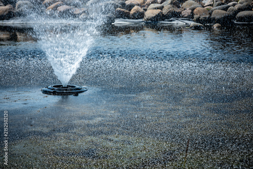 Obraz na plátně Floating aerating fountain spraying water droplets into shallow pond with a stone border landscaping background