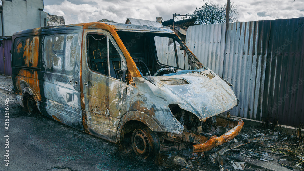 Obraz premium Side view of a completely burnt and destroyed delivery van in a war zone after military conflict, damaged fence background, war consequences concept