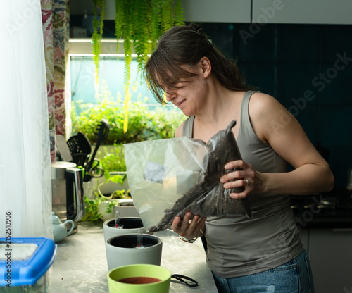 Woman in her 40s pouring soil from a bag into a flowerpot in a clean modern kitchen. Relaxed gardener filling and preparing substrate for repotting potted plant with happy calm enjoyment at home.