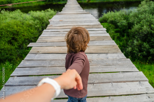 Caring mother holds her little boys hand while guiding young explorer across scenic wooden bridge in nature. This moment captures deep trust and parental protection as they discover outdoors together