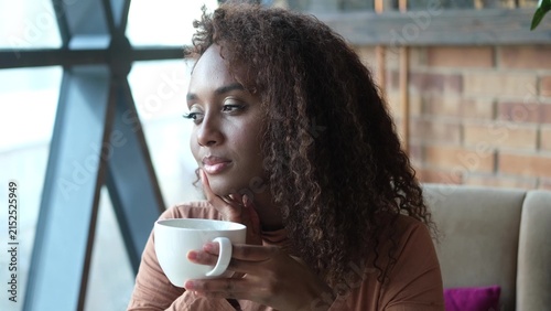 Bild auf Leinwand A very beautiful African American woman is sitting at a cafe table with a cup of coffee or tea