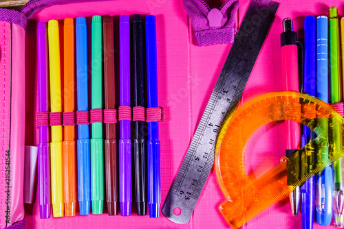 Different school stationeries (pens, pencils, felt tip pens, ruler and protractor) in a pink pencil box. Top view