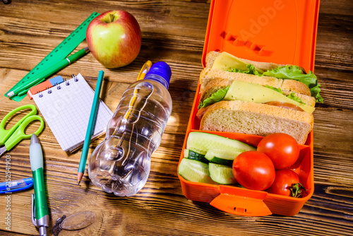 Bottle of water, ripe apple, different stationeries and lunch box with sandwiches, cucumbers and tomatoes on wooden table