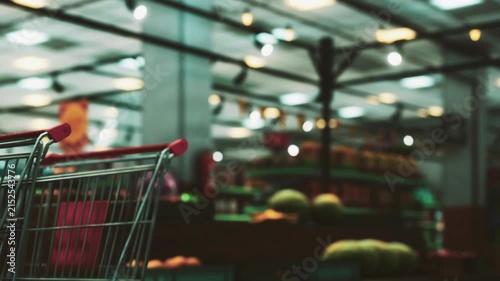 A shopping cart stands empty in a grocery store. Behind it, shelves display various fruits and vegetables. Customers walk around, selecting items for their meals.