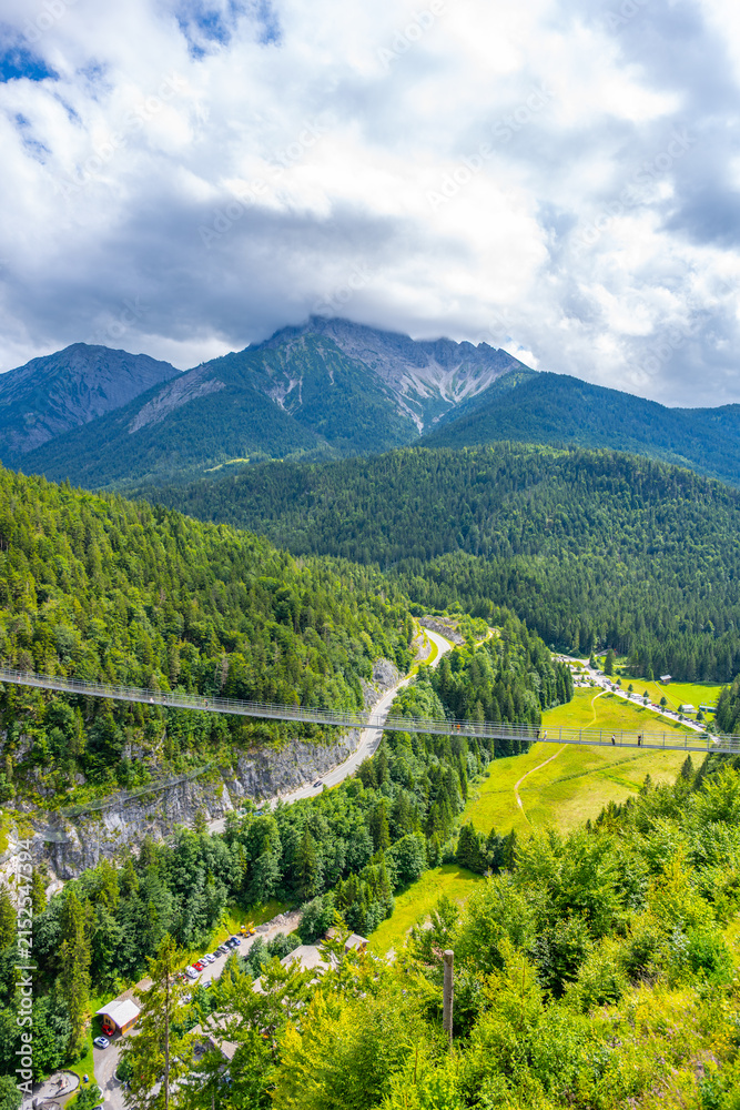 Obraz premium Highline 179 connects castle ruins with a Roman fortress. Visitors walk across the bridge over a wide river gorge. The scene shows mountain views and green landscape under a cloudy sky.