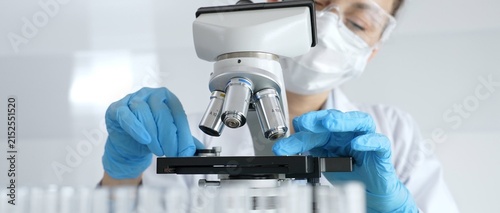 Woman science researcher wearing protective gear is checking microscope, analyzing blue test tubes in modern laboratory research setting. Medicine, and science concept