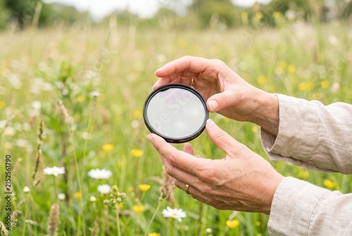 Hands holding a circular polarizing camera filter over a summer meadow with wild flowers