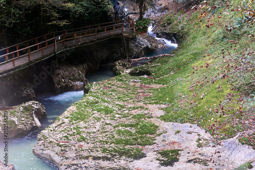 Wooden pedestrian bridge over a mountain river with turquoise water. Rocky bank covered with green moss and fallen leaves. Forest gorge, hiking trail.