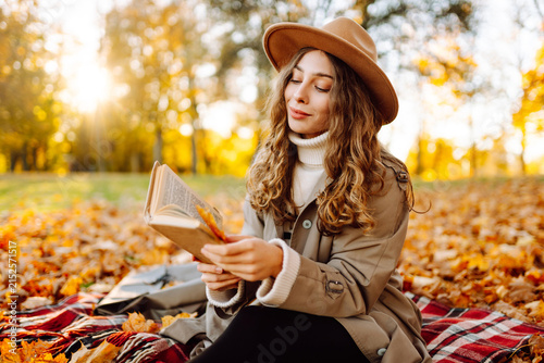 Woman in hat and coat sits on blanket in autumn park with book on sunny day. Beautiful woman reads book and relaxes surrounded by fallen leaves. Concept is autumn, relaxation, and enjoyment.
