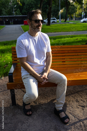 A middle-aged man in a white T-shirt and sunglasses stands on a street in the city. Portrait of a man with a beard