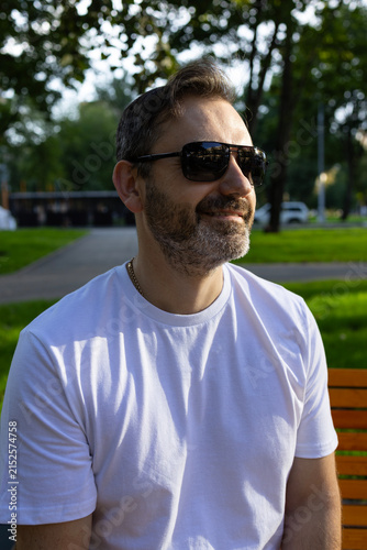 A middle-aged man in a white T-shirt and sunglasses stands on a street in the city. Portrait of a man with a beard