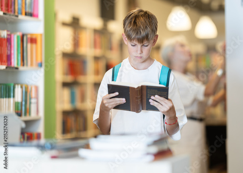 Canvas Print Interested teen male visitor holding a book near display stand in public library