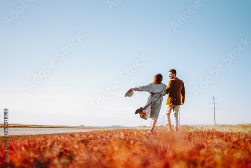 Young man and woman walk together across bright grassy field at sunset. A loving couple in stylish clothing enjoys nature and time together outdoors. Concept of love and relaxation.