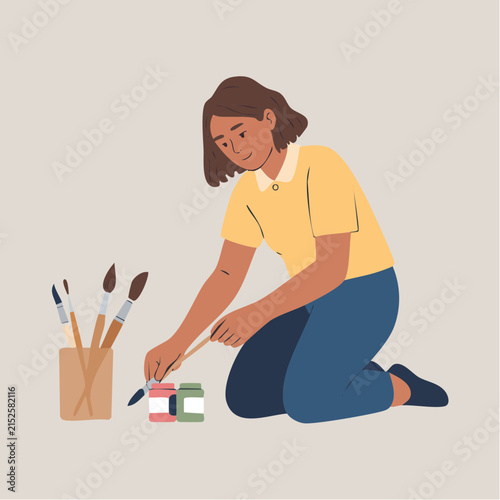 A young woman kneels on the floor, focused on painting with vibrant colors and brushes, capturing a moment of creative expression.