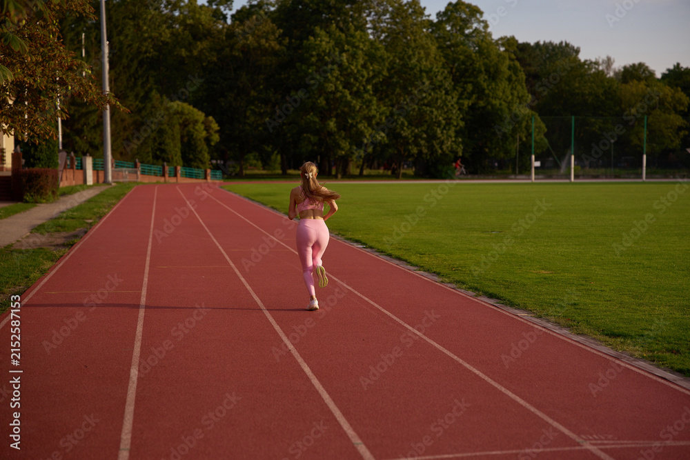 Fototapeta premium Active outdoor fitness, physical exercise, and athletic wellness concept. Young woman sprinting on a sports track. Female runner in motion during beautiful golden hour sunlight