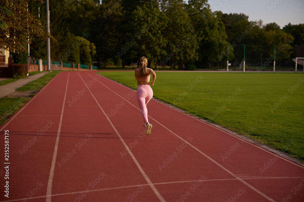 Obraz premium Female runner in motion during beautiful golden hour sunlight. Active outdoor fitness, physical exercise, and athletic wellness concept. Young woman sprinting on a sports track