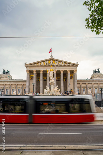 Austrian Parliament building in Vienna with red and white tram passing in front. Neoclassical architecture with golden sculptures, flag, and columns visible on overcast day.