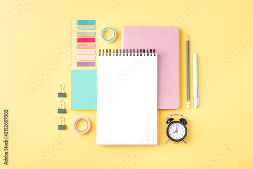 Stationery composition with blank notebook, ruler, paper clips, pencil and alarm clock on yellow surface. Back to school concept. Top view, flat lay, mockup