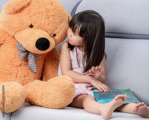Canvas Print A giant teddy bear accompanies an Asian little girl as she reads on the sofa, a heartwarming scene of family learning and childhood ideals
