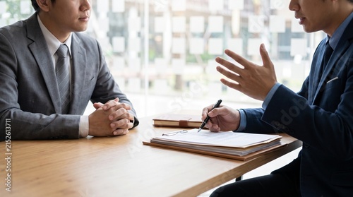 Two business professionals in suits discussing documents at a table, with one gesturing during the conversation