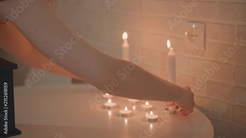 Close-up of woman hand lowering lit tealight onto glossy white tabletop beside scattered tealights and two pillar candles against painted brick wall, creating warm calm mood, with candle, tealight