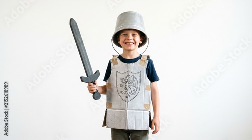 A young boy dressed as a knight, holding a toy sword and wearing a shield on his chest and helmet, smiles confidently in front of a white background.