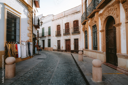 Winding cobblestone street in the historic center of Ronda, Spain, showing traditional Andalusian architecture with white walls, ochre details, and a shop displaying textiles