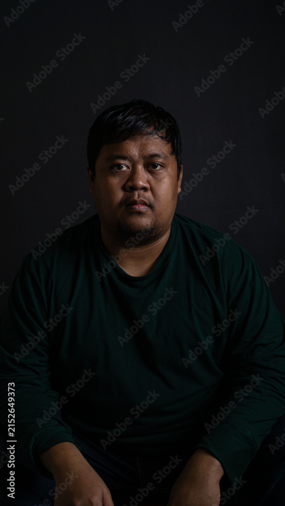 Fototapeta premium A compelling medium shot portrait of a confident Southeast Asian male sitting and looking directly into the camera against a solid dark gray background. The individual is wearing a