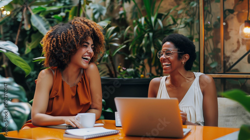 Two cheerful women collaborating with laptop in cozy modern workspace filled with plants