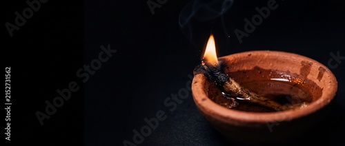 Traditional clay lamp with burning cotton wick and oil casting soft smoke against dark black background in cinematic atmosphere.