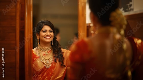 Beautiful Smiling Indian Bride in Traditional Red Saree and Heavy Gold Jewelry Looking in Mirror