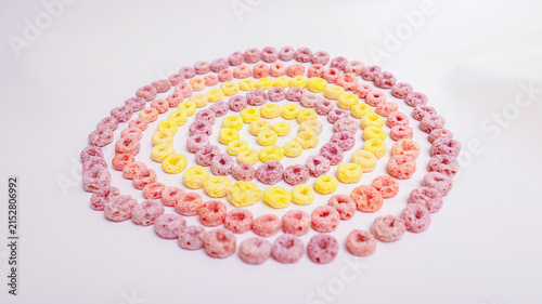 Colorful ring-shaped breakfast cereal in pink, yellow and orange arranged in a concentric spiral pattern on a white surface. Top view with copy space — perfect for breakfast, food, creativity and patt