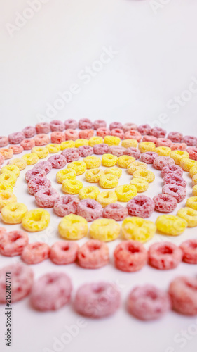 Colorful ring-shaped breakfast cereal in pink, yellow and orange arranged in a concentric spiral pattern on a white surface. Top view with copy space — perfect for breakfast, food, creativity and patt