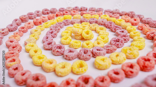 Colorful ring-shaped breakfast cereal in pink, yellow and orange arranged in a concentric spiral pattern on a white surface. Top view with copy space — perfect for breakfast, food, creativity and patt