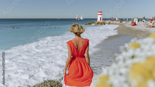 Woman in red dress walking along beach shore with waves crashing, sailboats in distance, and people relaxing under clear blue sky on sunny day