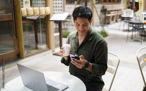 Young Asian man using a smartphone and drinking coffee while working at a cafe.