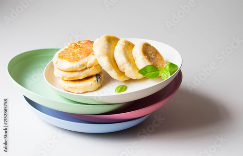 Homemade cottage cheese pancakes on the stack of flat multi-colored plates. Light background.