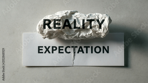 Reality versus Expectation: A crumpled piece of paper displaying the word REALITY sits atop a broken piece of paper with the word EXPECTATION printed on it.