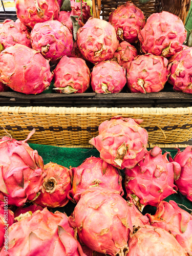 Bright pink dragon fruits pile high in woven baskets. Their scales glisten under warm market lighting. Vibrant colors draw attention to fresh, exotic produce. Ideal for food, health. No people present