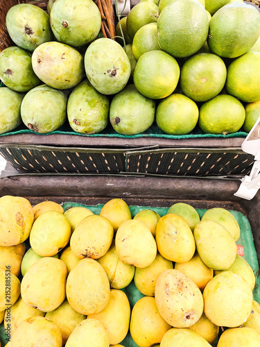 Fresh mangoes and lemons display vibrant colors. Bright yellow lemons sit beside green mangoes. Baskets hold fruit in a market stall. Natural sunlight highlights each fruit's texture
