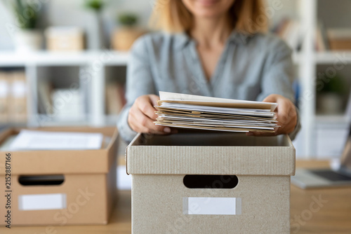 Woman holds papers above a cardboard box. She prepares to archive or discard documents. Background shows office shelves and another box. Her expression suggests focus or mild fatigue