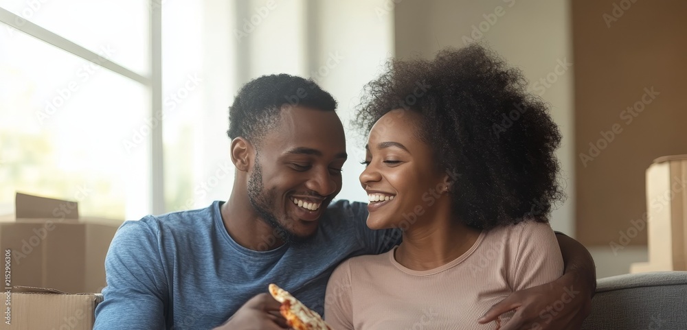 Obraz premium Happy young african american couple eating pizza together while sitting in their new home